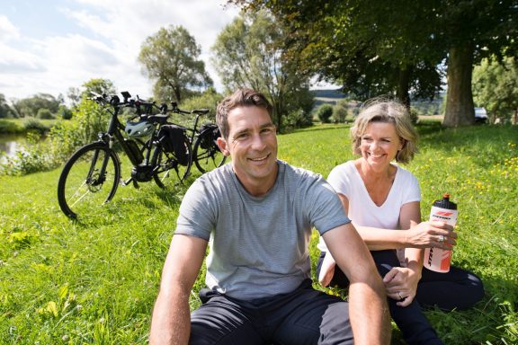 Zwei lächelnde Personen sitzen auf Grünfläche neben Fahrrädern und genießen Erfrischungen.