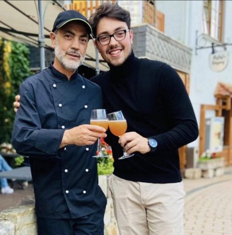 Zwei Männer, einer im Koch-Outfit, stoßen mit Getränken an. Hintergrund mit Pflanzen und Gebäuden.