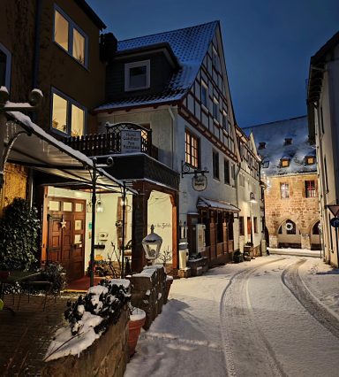 Schneebedeckte Straße mit traditionellen Gebäuden und beleuchteten Fenstern bei Nacht.