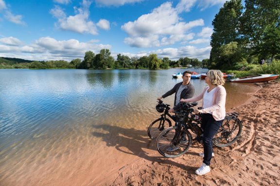 Zwei Personen mit Fahrrädern am Ufer eines ruhigen Sees, umgeben von Bäumen.