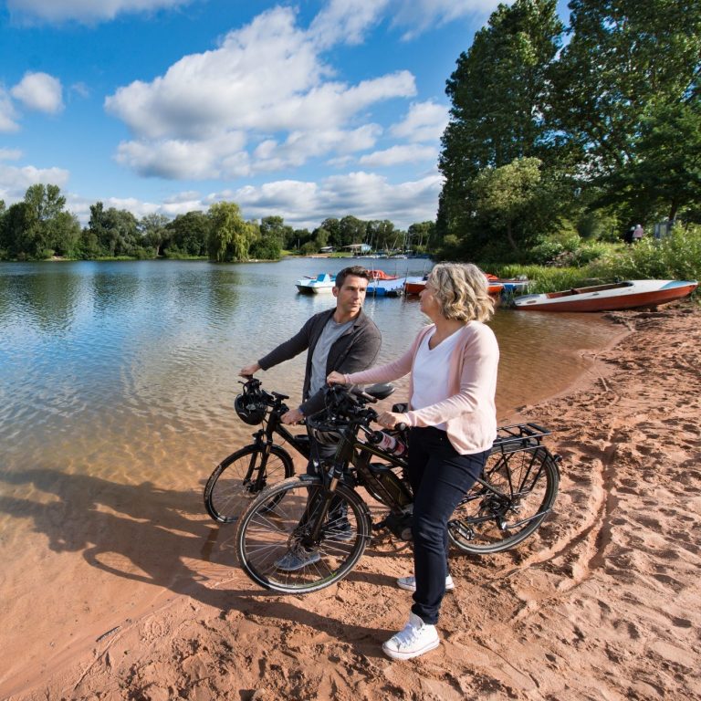 Zwei Personen mit Fahrrädern am Ufer eines ruhigen Sees. Boote im Hintergrund.