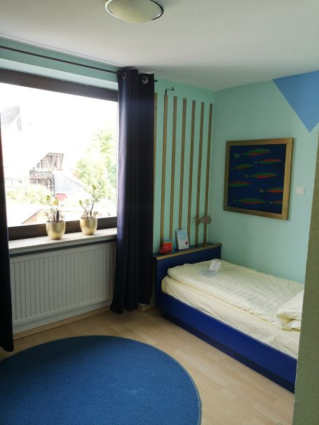 Ein helles Zimmer mit einem Bett, Teppich und Fenstern, umgeben von Pflanzen.