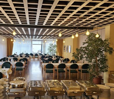 Modernes Restaurant mit Tischen, Pflanzen und Buffet-Wärmeschalen in einem hellen Raum.