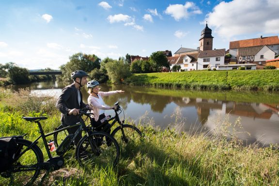 Ein Paar steht neben Fahrrädern am Fluss und zeigt auf eine Stadtansicht im Hintergrund.