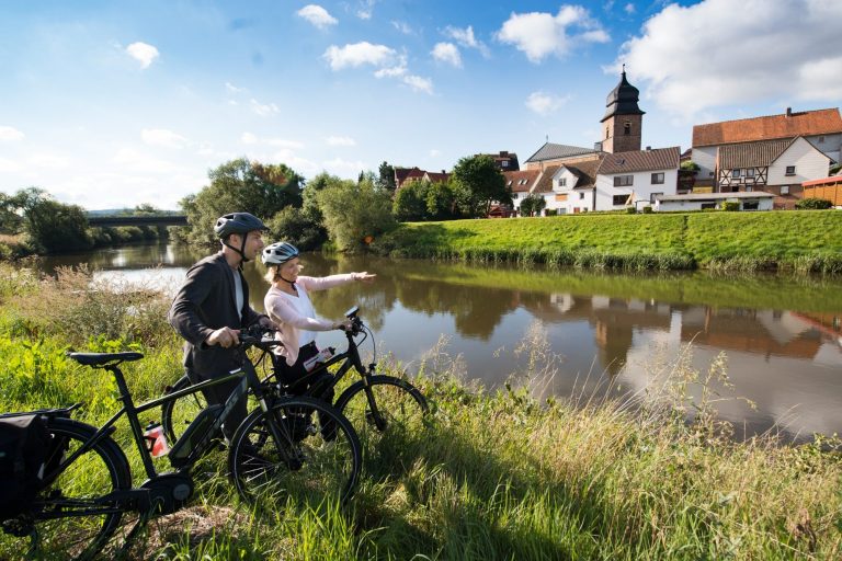 Paar mit Fahrrädern am Fluss, zeigt auf die Stadt im Hintergrund.