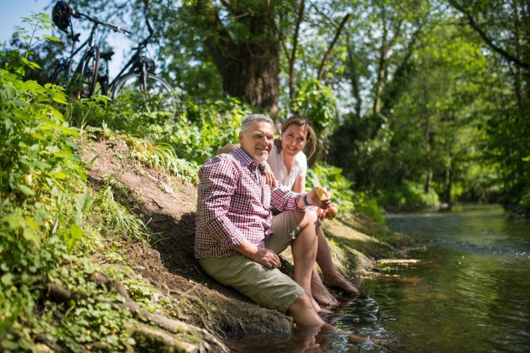 Älteres Paar sitzt am Ufer eines Baches und genießt die Natur. Fahrräder im Hintergrund.