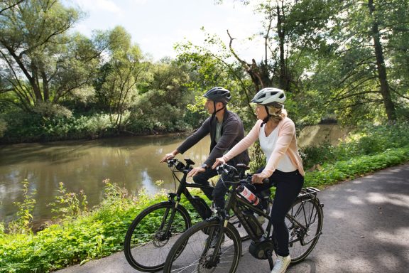 Zwei Radfahrer entlang eines Flusses, umgeben von grüner Vegetation.