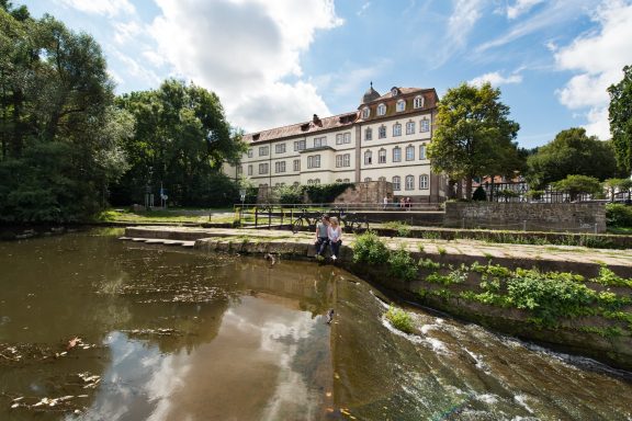 Historisches Gebäude am Ufer eines ruhigen Gewässers, umgeben von Bäumen.