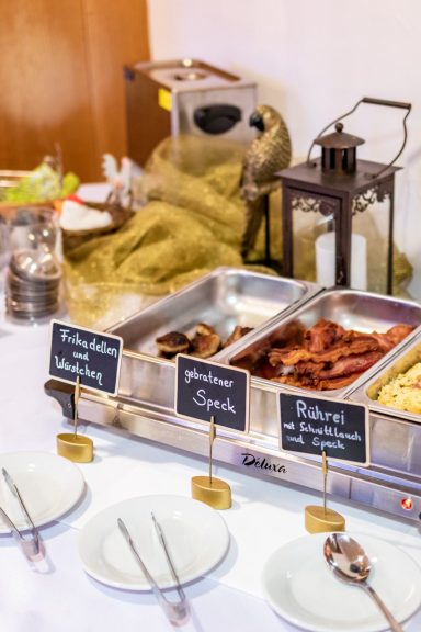 Buffet mit verschiedenen Gerichten, darunter Speck und Pasta, das auf einem Tisch angerichtet ist.