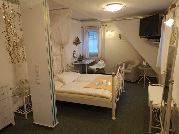 Helles Zimmer mit Doppelbett, Tisch, Stuhl und Fernseher. Gemütliche Sitzecke vorhanden.