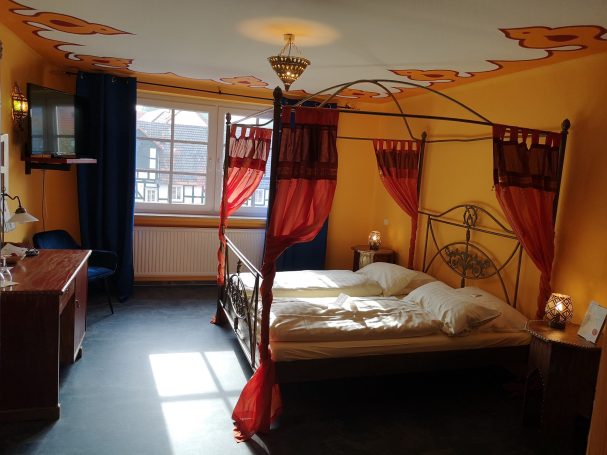 Helles Schlafzimmer mit Himmelbett, roten Vorhängen und blauer Dekoration.