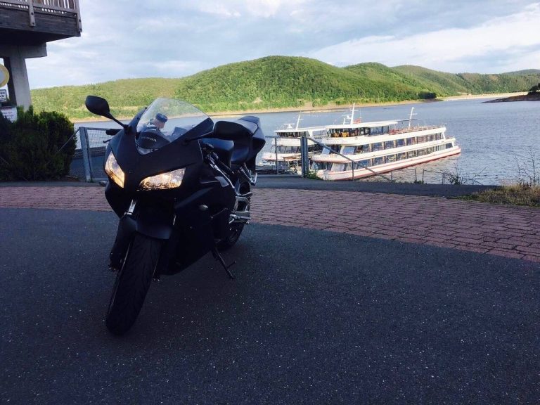 Schwarzes Motorrad vor einem See und grünen Hügeln. Im Hintergrund ein Boot.