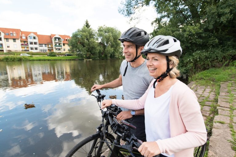 Paar mit Fahrrädern am Ufer eines ruhig fließenden Gewässers, lächelnd und in Freizeitkleidung.