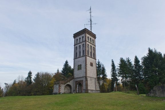Historischer Aussichtsturm auf einer Wiese, umgeben von Bäumen und blauem Himmel.
