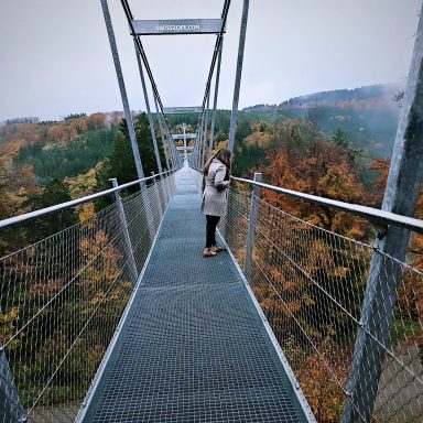 Frau auf einer Hängebrücke mit herbstlichen Bäumen im Hintergrund.