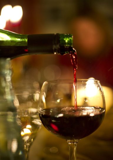 Rote Wein wird aus einer Flasche in ein Weinglas gegossen, im Hintergrund Kerzenlicht.