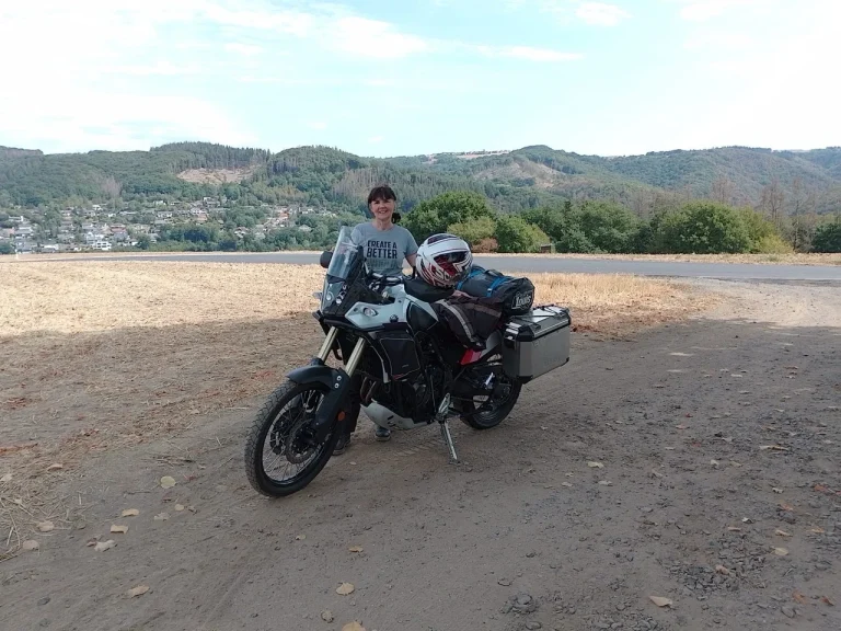 Zwei Personen stehen neben einem Motorrad auf sandigem Boden mit Bergen im Hintergrund.