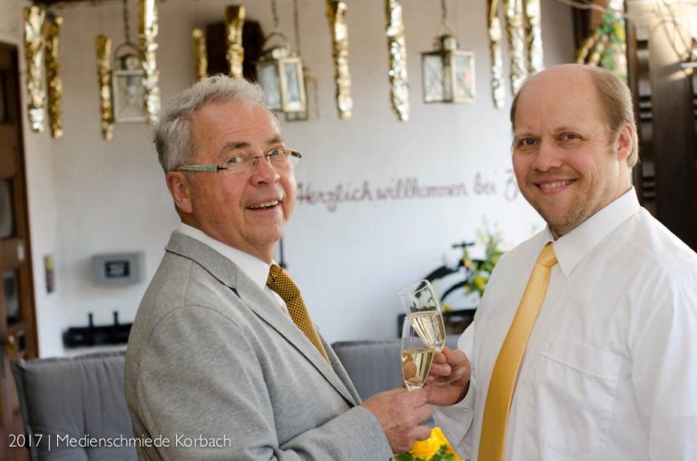 Zwei Männer in Anzügen lächeln und stoßen mit Gläsern an, festliche Dekoration im Hintergrund.
Inhaber Uli und Urs Zabel 