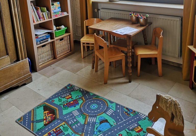 Kinderzimmer mit Tisch, Stühlen, Spielteppich und Bücherregal.