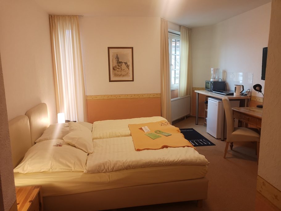 Heller Hotelraum mit Doppelbett, Tisch, Kühlschrank und Kaffeezubereiter.