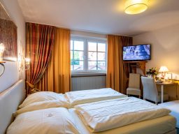 Helles Hotelzimmer mit zwei Betten, Schreibtisch und Fernseher. Gardinen in warmen Farben.