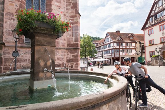 Brunnen in einer Straße mit Radfahrern und historischen Gebäuden im Hintergrund.