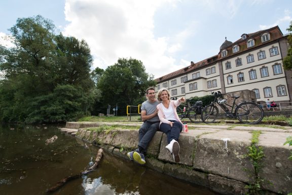 Zwei Personen sitzen am Ufer eines Gewässers, im Hintergrund Fahrräder und ein Gebäude.