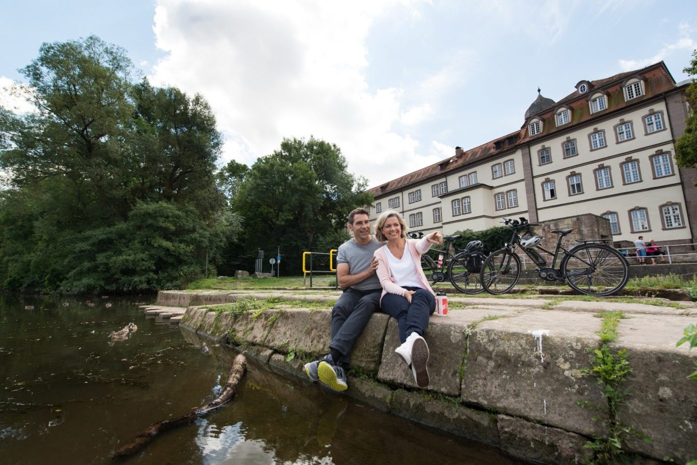 Paar sitzt am Ufer eines Gewässers mit Fahrrädern im Hintergrund und lächelt.