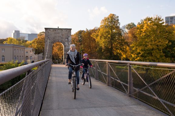 Zwei Radfahrer, ein Erwachsener und ein Kind, fahren über eine Brücke im herbstlichen Park.