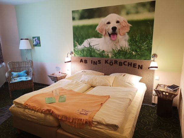 Helles Zimmer mit Bett, Tisch und Wandbild eines lächelnden Welpen auf Gras.