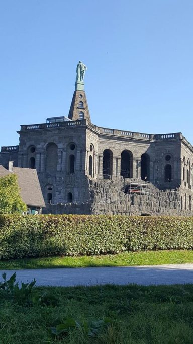 Historisches Gebäude mit Statue auf dem Dach, umgeben von Grünflächen.