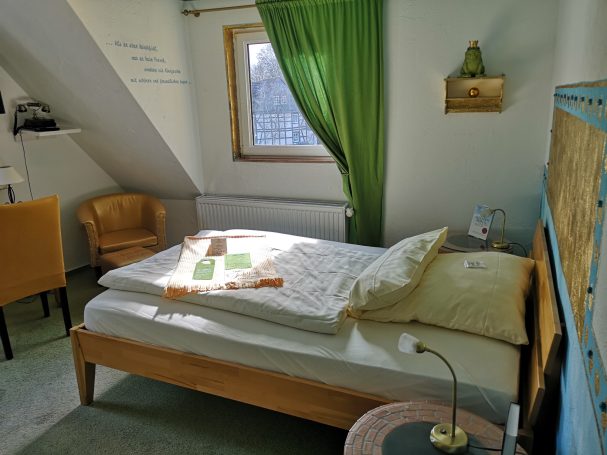 Helles Schlafzimmer mit Bett, Stuhl, Fenster, grünen Vorhängen und Nachttischlampe.