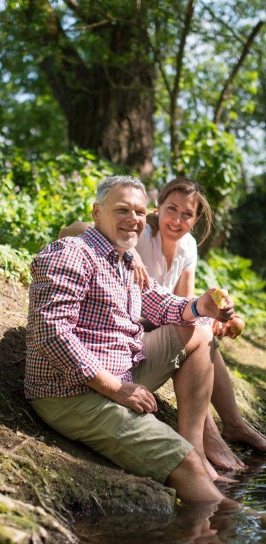 Senior Mann und junge Frau sitzen am Ufer eines Flusses, umgeben von Bäumen und Grün.