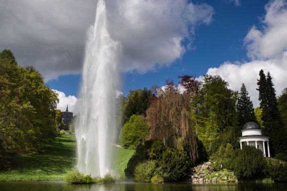 Ein sprudelnder Wasserfall in einem grünen Park mit Bäumen und einem Pavillon.