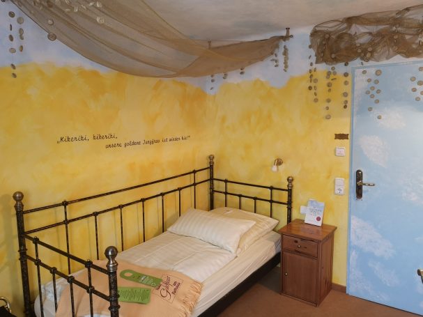 Schlafzimmer mit einem Bett, gelber Wandfarbe und Holzdekor, dazu ein Nachttisch und Tür.