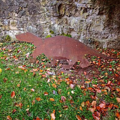 Eisenfigur eines Dinosauriers vor einer Steinmauer, umgeben von buntem Laub.