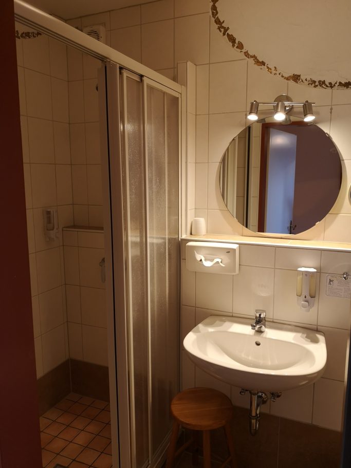 Badezimmer mit Dusche, Waschbecken und runder Spiegelleuchte.